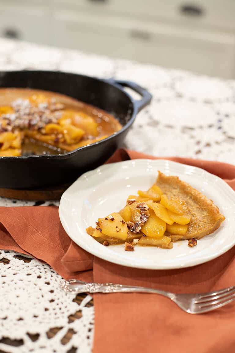 A slice of sourdough Dutch baby pancake topped with caramelized apples and chopped pecans on a white plate, set on a burnt orange napkin. In the background, a cast-iron skillet with the remaining pancake sits on a lace-covered countertop.