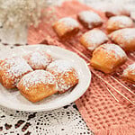 Plate of freshly fried sourdough beignets dusted with powdered sugar with more beignets cooling on a rack behind it. 2.