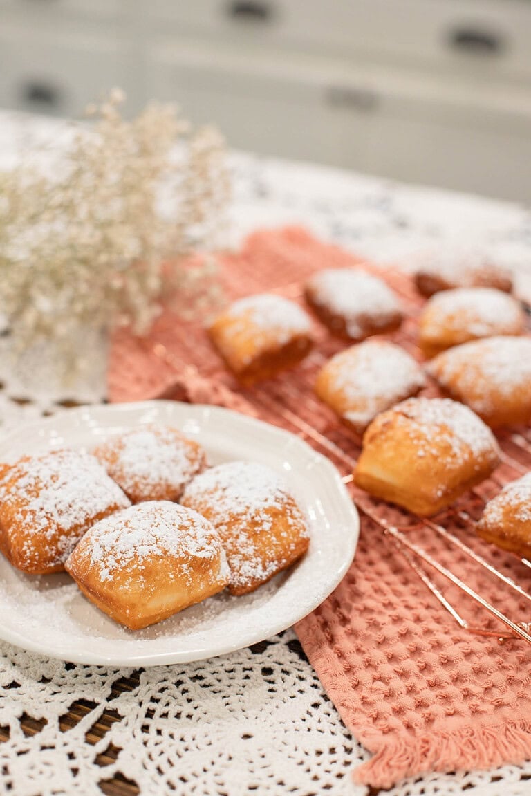 Plate of freshly fried sourdough beignets dusted with powdered sugar with more beignets cooling on a rack behind it. 2.