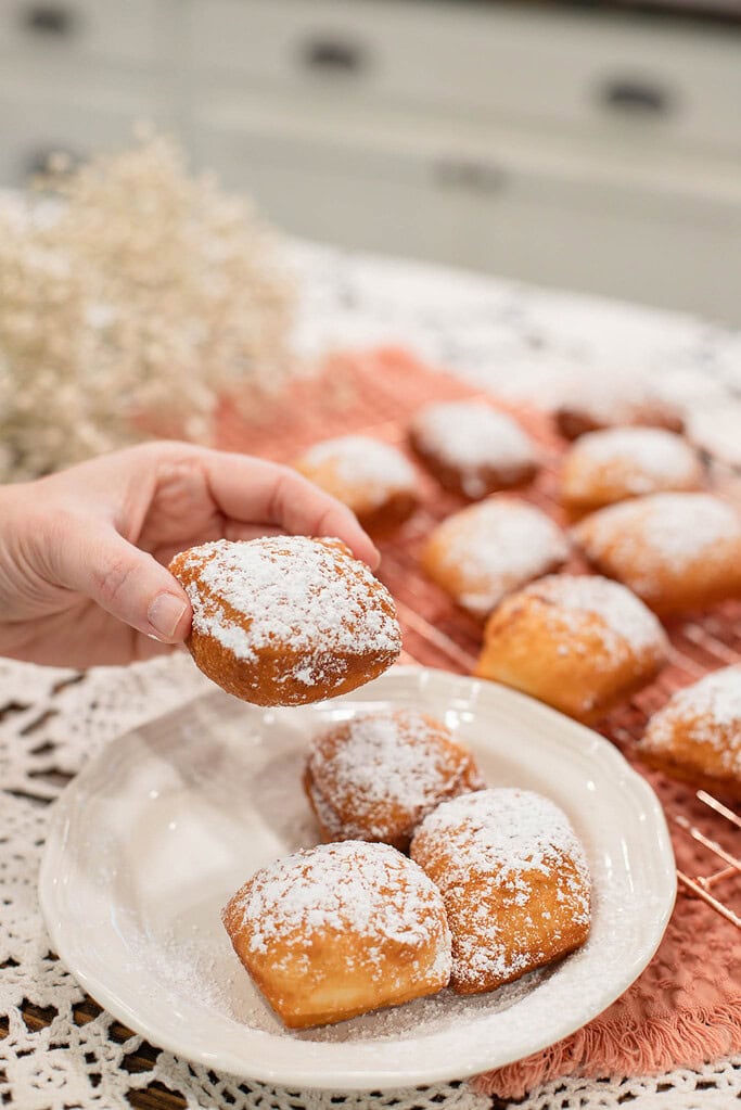 Hand picking up a warm sourdough beignet covered in powdered sugar from a plate