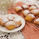 Golden, pillowy sourdough beignets dusted with powdered sugar rest on a cream-colored plate, with more beignets cooling on a copper wire rack in the background. A warm, textured napkin and delicate baby’s breath flowers add a cozy, cottage-inspired touch to the scene.