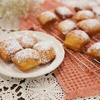 Golden, pillowy sourdough beignets dusted with powdered sugar rest on a cream-colored plate, with more beignets cooling on a copper wire rack in the background. A warm, textured napkin and delicate baby’s breath flowers add a cozy, cottage-inspired touch to the scene.