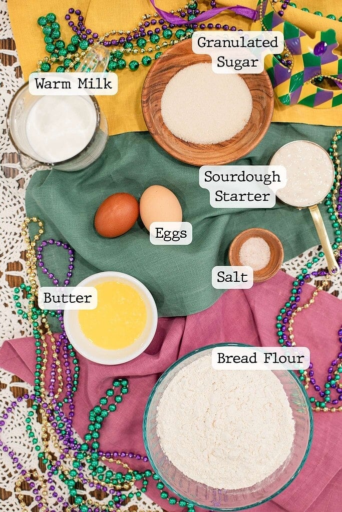 Flat lay of sourdough king cake dough ingredients including warm milk, sugar, sourdough starter, eggs, butter, salt, and bread flour