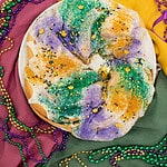 A fully baked sourdough king cake covered in white icing, topped with green, purple, and gold sugar sprinkles, and decorated with small festive sprinkles. Mardi Gras beads and a harlequin-style mask are scattered around.