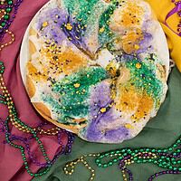A fully baked sourdough king cake covered in white icing, topped with green, purple, and gold sugar sprinkles, and decorated with small festive sprinkles. Mardi Gras beads and a harlequin-style mask are scattered around.