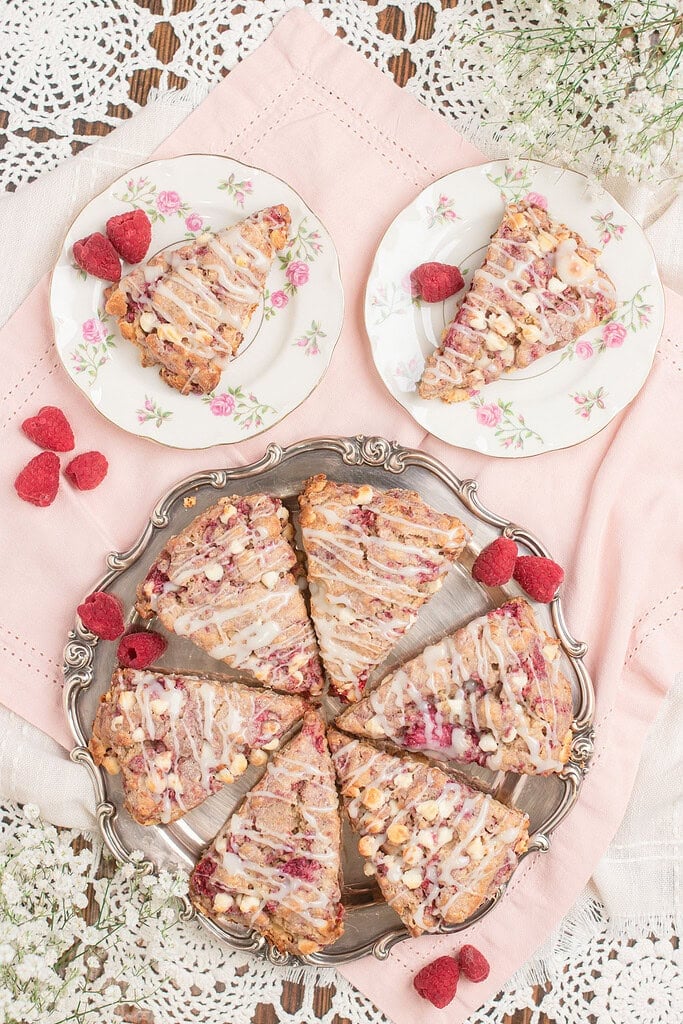 Overhead flat lay of sourdough raspberry white chocolate scones with vanilla glaze drizzle arranged on a silver tray and two floral china plates, surrounded by fresh raspberries and baby's breath on a pink linen