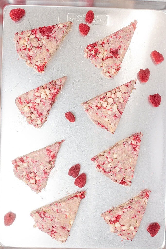 Eight unbaked sourdough raspberry white chocolate scone wedges arranged on a silver baking sheet, scattered with fresh raspberries