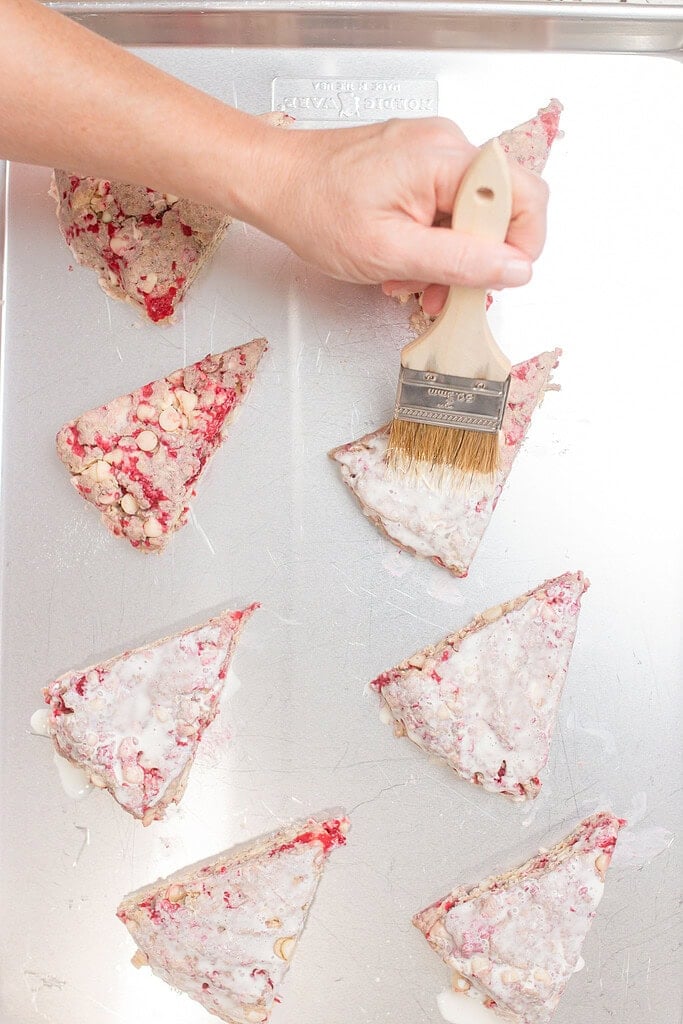 Hand brushing heavy cream onto unbaked raspberry white chocolate scone wedges on a silver baking sheet before baking