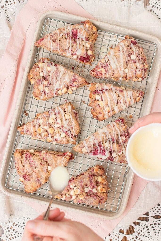 Overhead view of baked sourdough raspberry white chocolate scones on a wire rack set in a baking pan, with a hand spooning vanilla glaze from a white bowl
