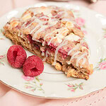 Close-up of a glazed sourdough raspberry white chocolate scone on a floral china plate with two fresh raspberries, silver tray of scones blurred in the background