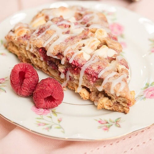 Close-up of a glazed sourdough raspberry white chocolate scone on a floral china plate with two fresh raspberries, silver tray of scones blurred in the background