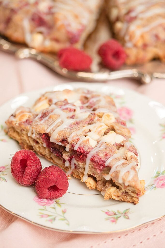 Close-up of a glazed sourdough raspberry white chocolate scone on a floral china plate with two fresh raspberries, silver tray of scones blurred in the background