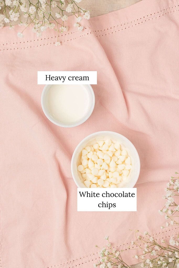 Flat lay of two white ramekins on a pink linen -- one filled with cream and one with white chocolate chips -- surrounded by sprigs of baby's breath