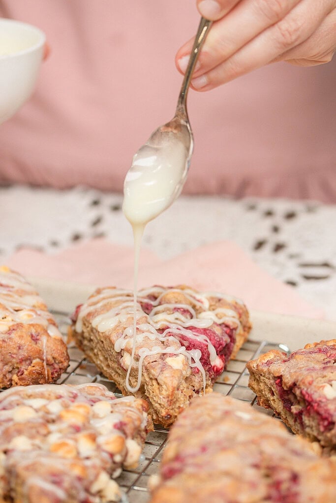 Close-up of a hand drizzling vanilla glaze from a spoon over freshly baked sourdough raspberry white chocolate scones cooling on a wire rack