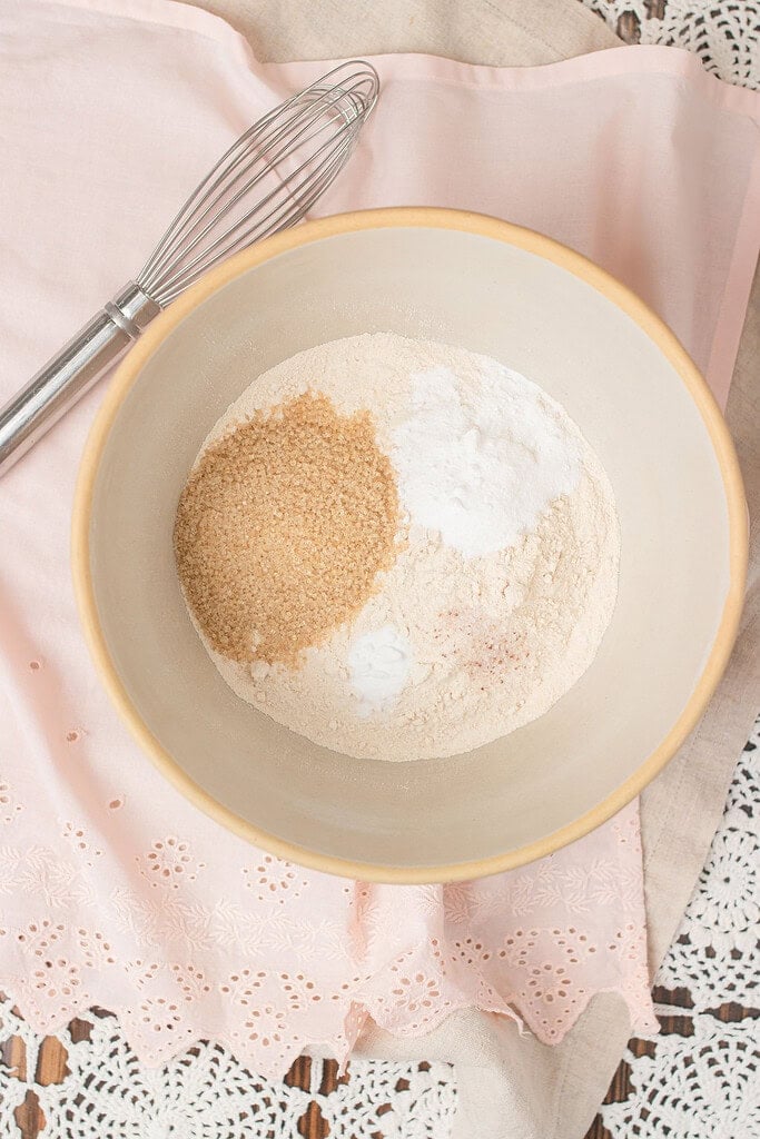 Overhead view of freshly milled whole wheat flour, sugar, and baking powder measured into a gray mixing bowl beside a wire whisk on a pink linen