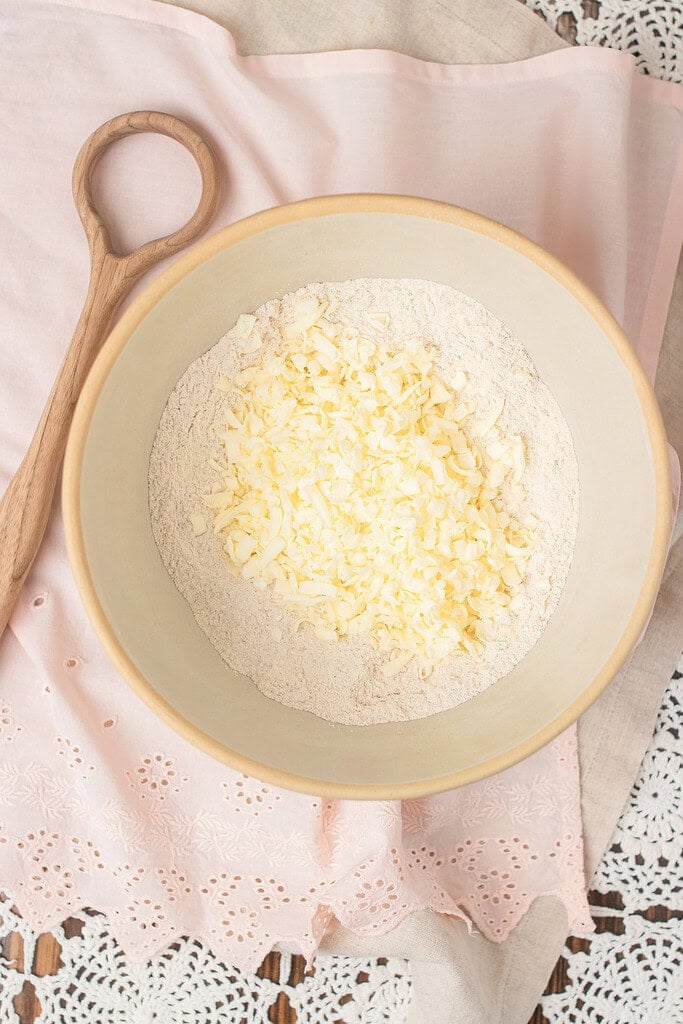 Overhead view of grated cold butter scattered over freshly milled flour in a gray mixing bowl, with a wooden spoon and pink linen beside it