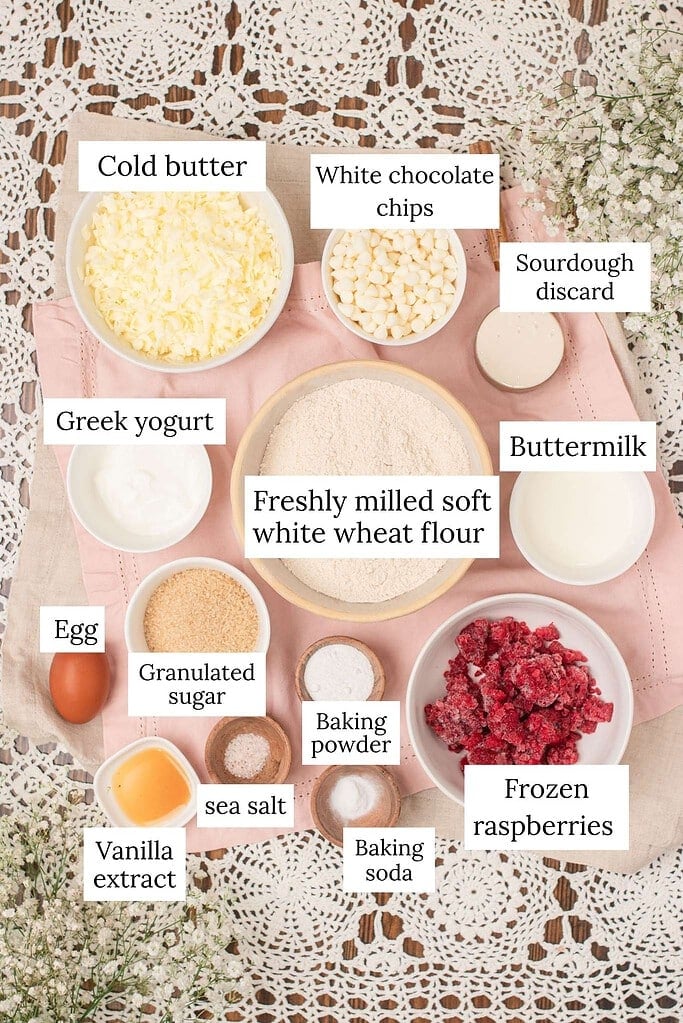 overhead ingredient flat lay for sourdough raspberry white chocolate scones showing freshly milled soft white wheat flour, cold grated butter, white chocolate chips, sourdough discard, Greek yogurt, buttermilk, egg, granulated sugar, frozen raspberries, baking powder, baking soda, sea salt, and vanilla extract in bowls on a pink eyelet linen with crochet lace and white baby's breath.