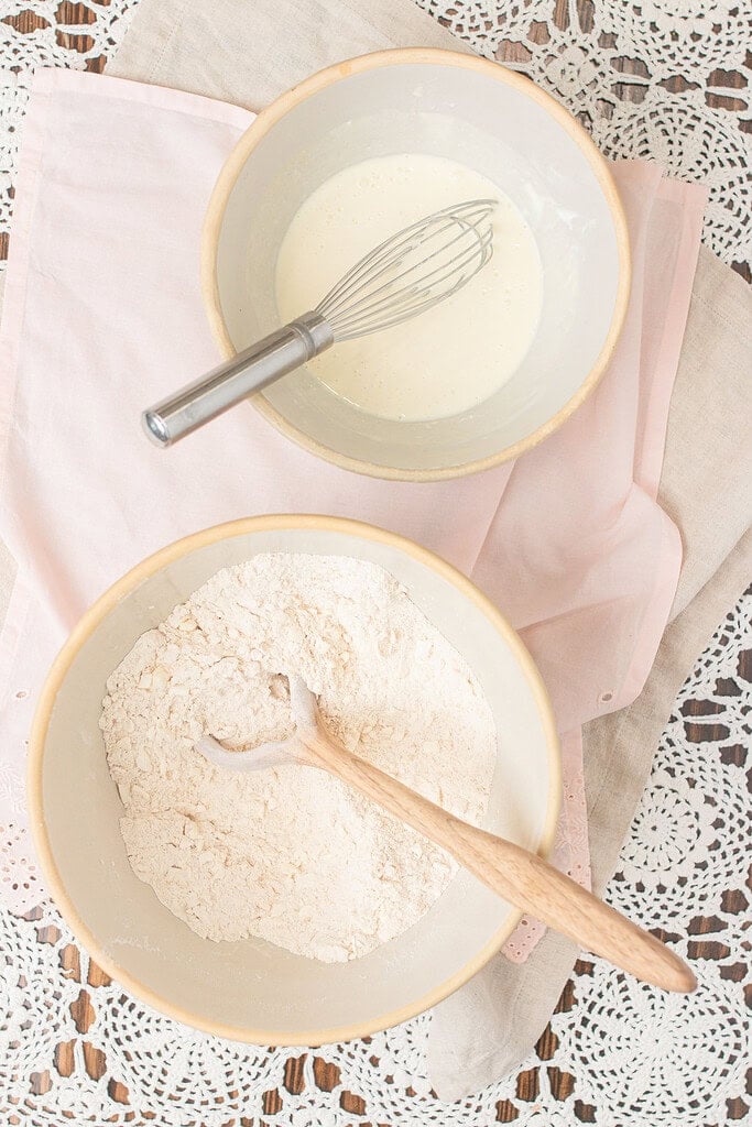 Overhead view of two mixing bowls on a pink linen -- one with freshly milled flour and a wooden spoon, one with a cream mixture and a whisk