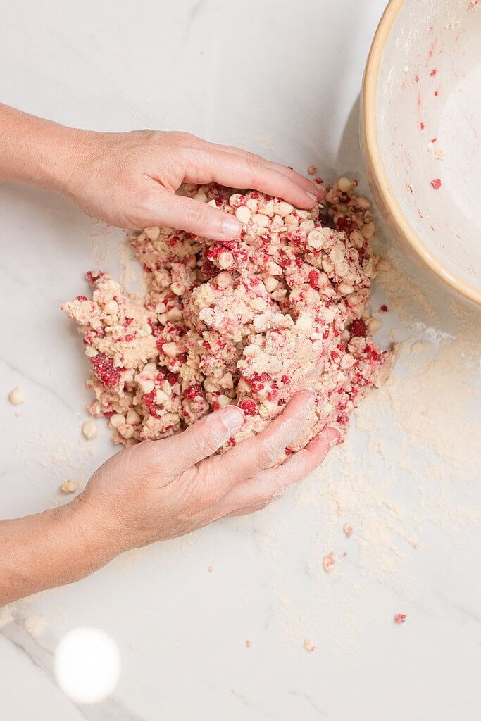 Hands pressing together shaggy sourdough scone dough studded with raspberries and white chocolate chips on a floured marble surface