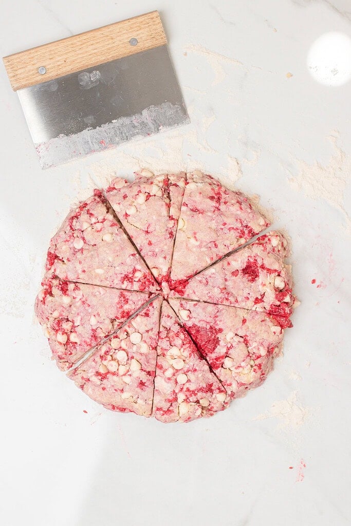 Overhead view of sourdough raspberry white chocolate scone dough shaped into a round disc and cut into eight wedges on a marble surface, with a bench scraper nearby