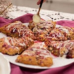 A hand lifting a glazed raspberry white chocolate sourdough scone from a plate, highlighting its texture and layers.