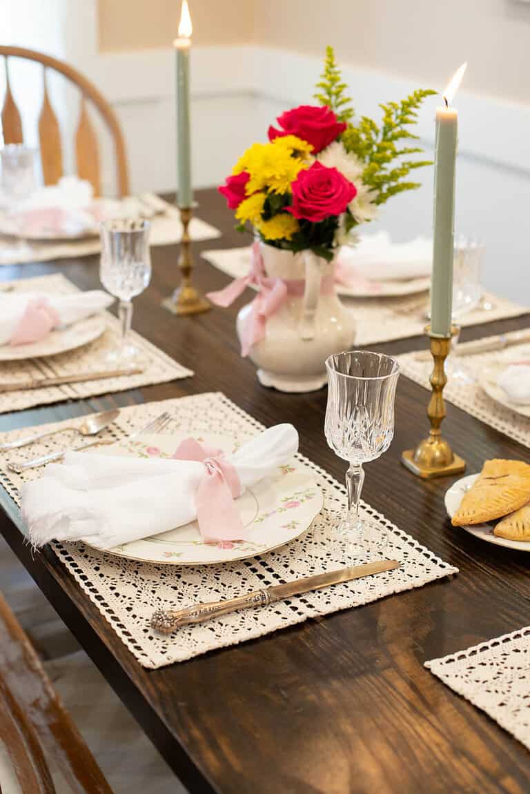Elegant cottage-style spring tablescape with vintage floral plates, delicate lace placemats, and soft pink ribbon-tied napkins. A glowing candle and fresh floral centerpiece add charm.