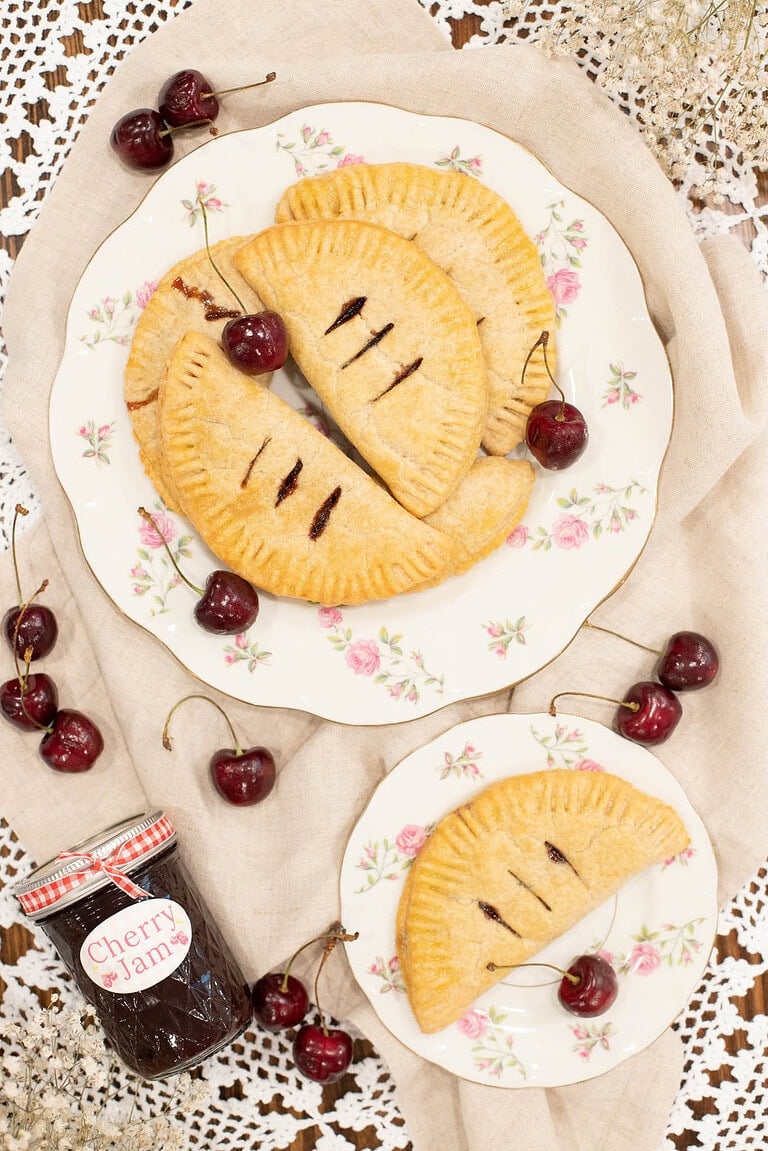 Cherry hand pies on floral plates with fresh cherries and homemade cherry jam on a lace tablecloth