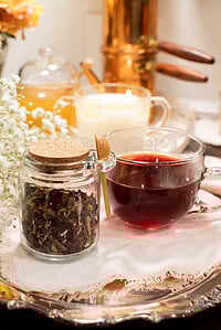 Glass jar of loose cherry tea blend with cork lid beside a clear glass cup filled with deep red cherry tea on a silver tray. A lit candle, honey jar, and copper kettle sit in the cozy background with white flowers.