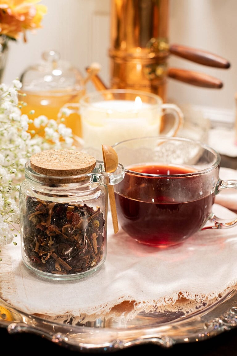 Glass jar of loose cherry tea blend with cork lid beside a clear glass cup filled with deep red cherry tea on a silver tray. A lit candle, honey jar, and copper kettle sit in the cozy background with white flowers.
