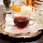 Clear glass teacup filled with deep red cherry tea placed on a vintage silver tray with baby’s breath flowers and cozy cottage kitchen decor behind.