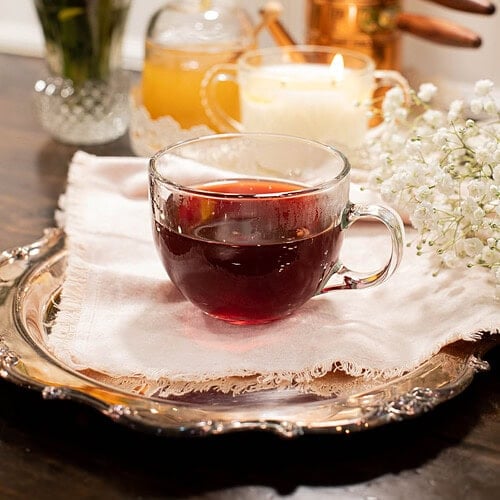 Clear glass teacup filled with deep red cherry tea placed on a vintage silver tray with baby’s breath flowers and cozy cottage kitchen decor behind.