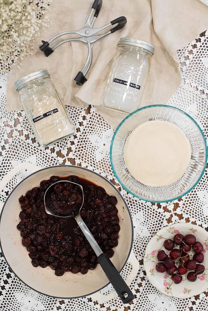 A flat-lay of jam-making ingredients on a lace-covered table, including a pot of cooked cherries, a bowl of sugar, a jar of calcium water, a jar of Pomona’s pectin, a jar lifter, and a small plate of fresh cherries.