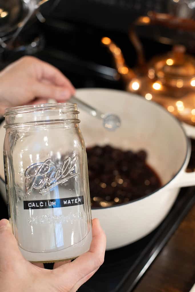 A person holds a glass Ball jar labeled “Calcium Water” while using a measuring spoon to add an ingredient to a white Dutch oven filled with simmering cherries. A copper kettle sits in the background on the stovetop.