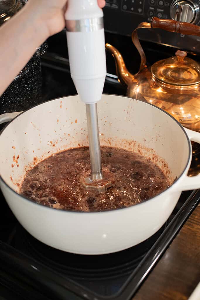An immersion blender is being used to blend the cooked cherry mixture in a white Dutch oven. The dark red jam is bubbling, and a copper kettle is visible in the background.