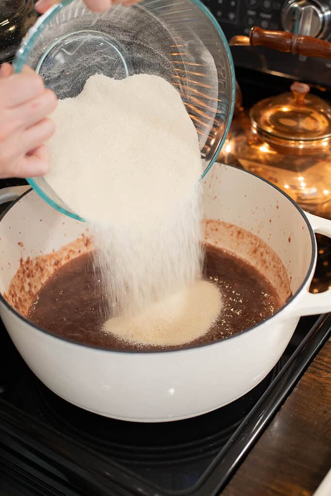 A person pours a large bowl of granulated sugar into the blended cherry mixture in a white Dutch oven. The sugar cascades into the bubbling jam.