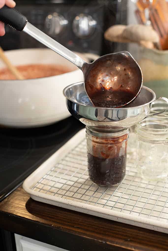 A person ladles hot cherry jam into a sterilized mason jar through a stainless steel canning funnel, which is placed over a cooling rack on a baking sheet.
