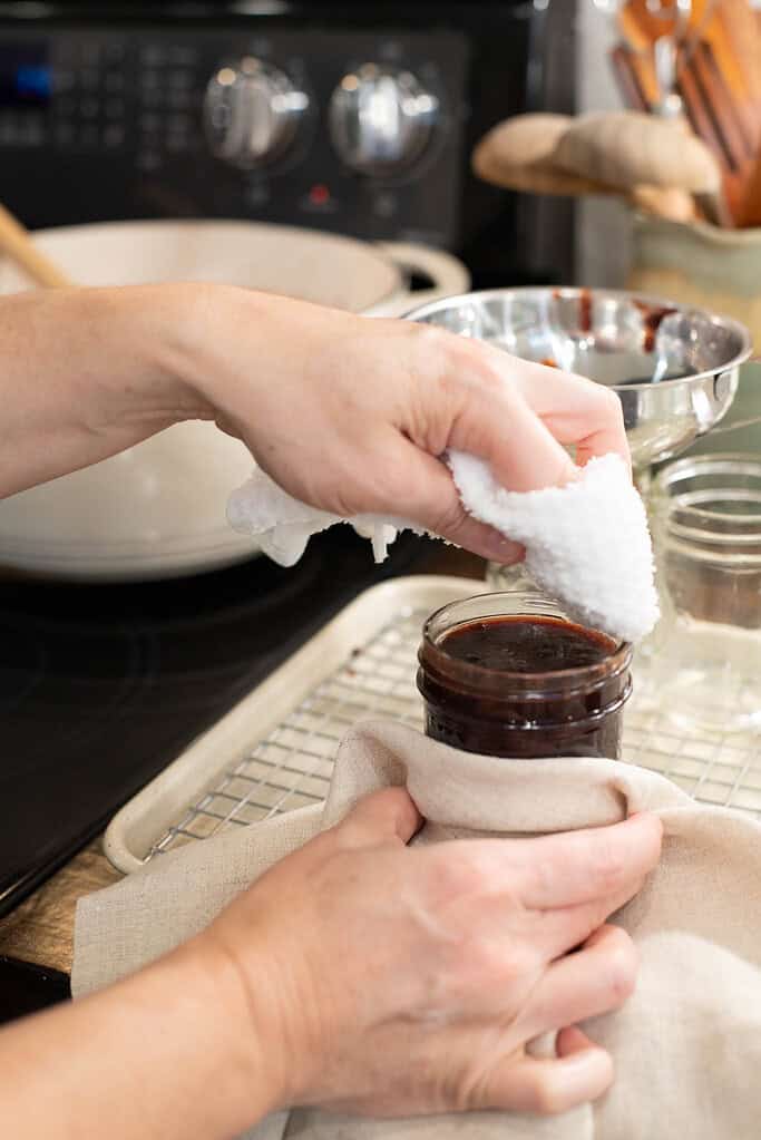 A person wipes the rim of a filled mason jar with a clean cloth, preparing it for sealing. The jam-filled jar sits on a cooling rack.