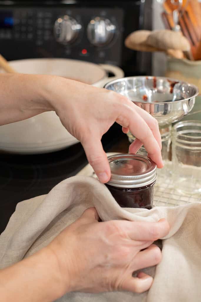 A person screws a canning ring onto the jar, using a cloth for grip, ensuring it is fingertip-tight.