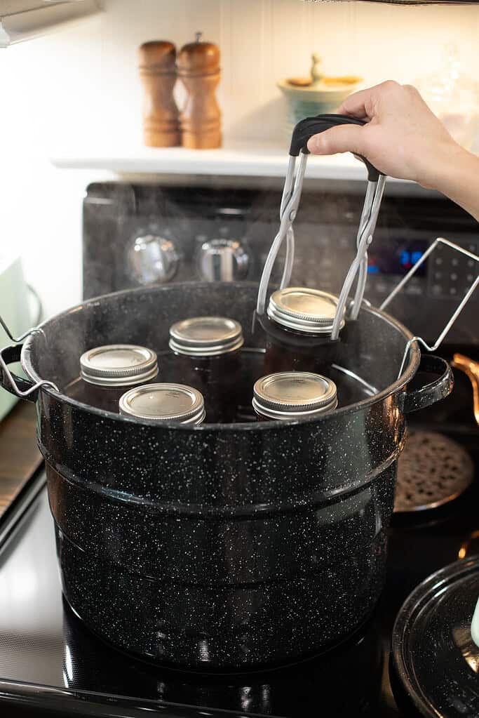 A person uses a jar lifter to place sealed mason jars of cherry jam into a boiling water canner for processing.