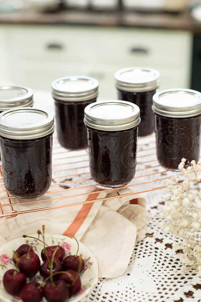 A finished batch of homemade cherry jam cools on a copper wire rack, with six sealed jars arranged neatly. A plate of fresh cherries and a linen cloth add a rustic touch.