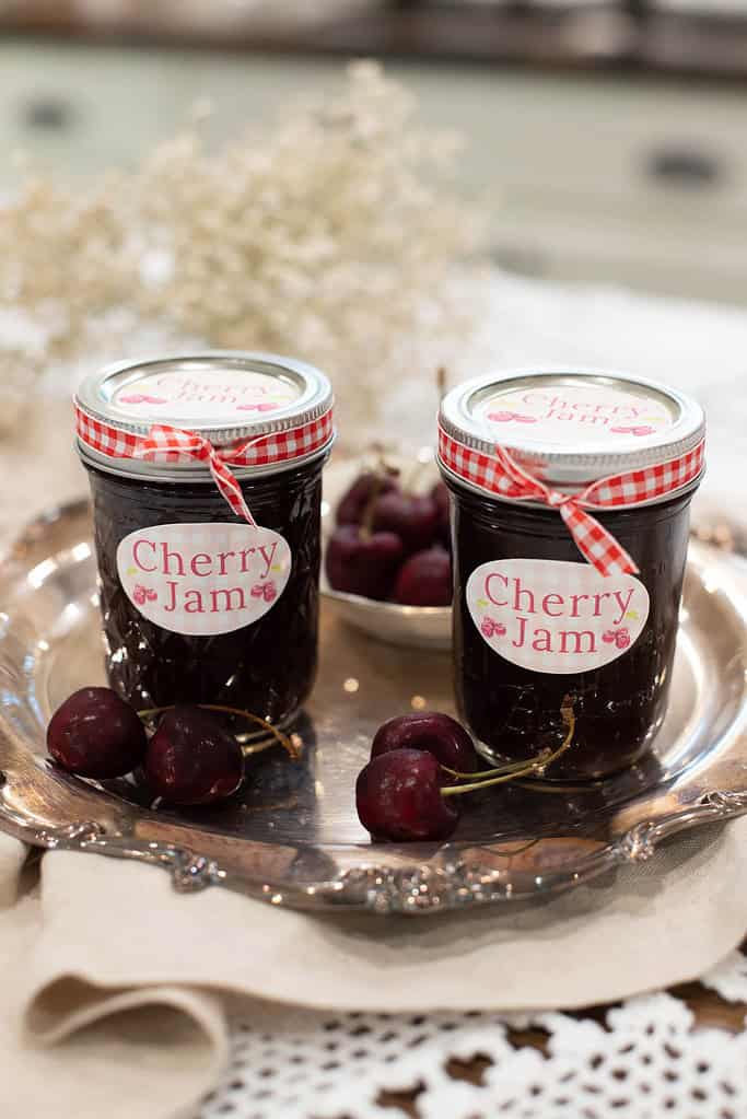 A close-up of two cherry jam jars on a silver tray, decorated with red and white gingham ribbon and classic labels, with cherries scattered around.
