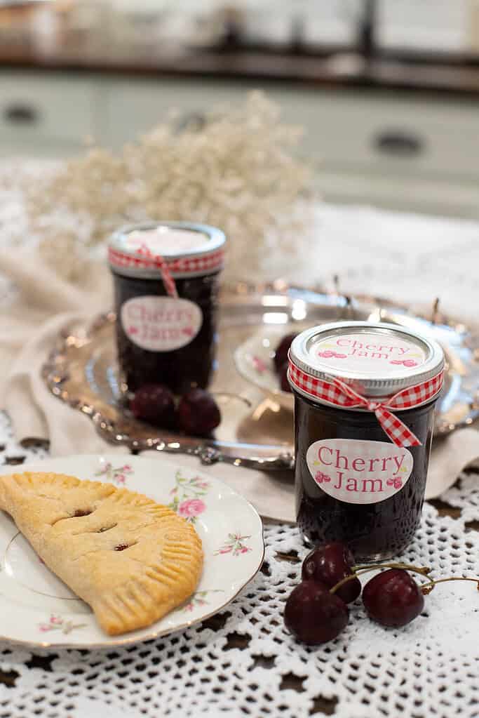 A plate of golden, flaky cherry hand pie sits next to a jar of cherry jam on a lace tablecloth, with fresh cherries and vintage dishware.