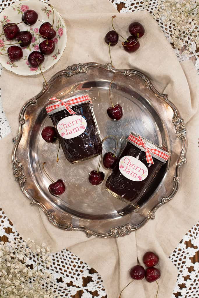 A flat-lay of two cherry jam jars resting on a vintage silver tray, surrounded by fresh cherries, a linen cloth, and lace decor.