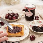 A slice of golden toast being spread with cherry jam, served on vintage floral dishware, with a bowl of fresh cherries.