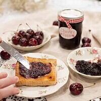 A slice of golden toast being spread with cherry jam, served on vintage floral dishware, with a bowl of fresh cherries.