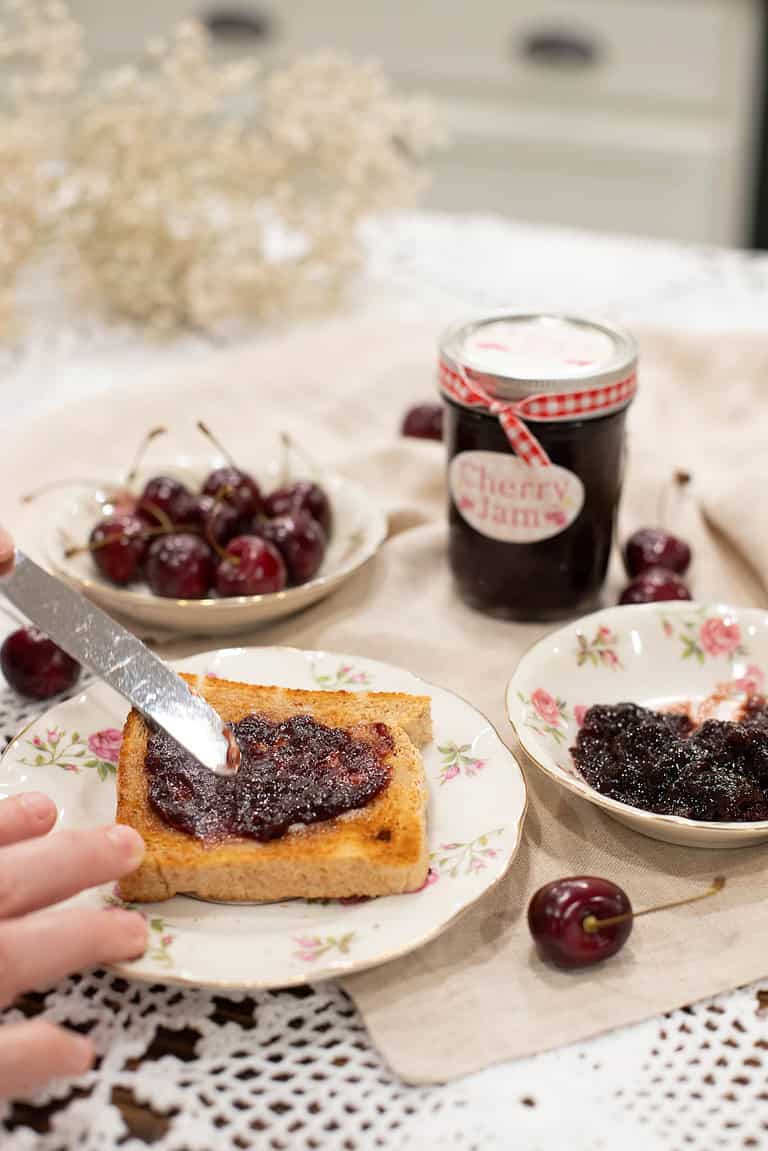 A slice of golden toast being spread with cherry jam, served on vintage floral dishware, with a bowl of fresh cherries.