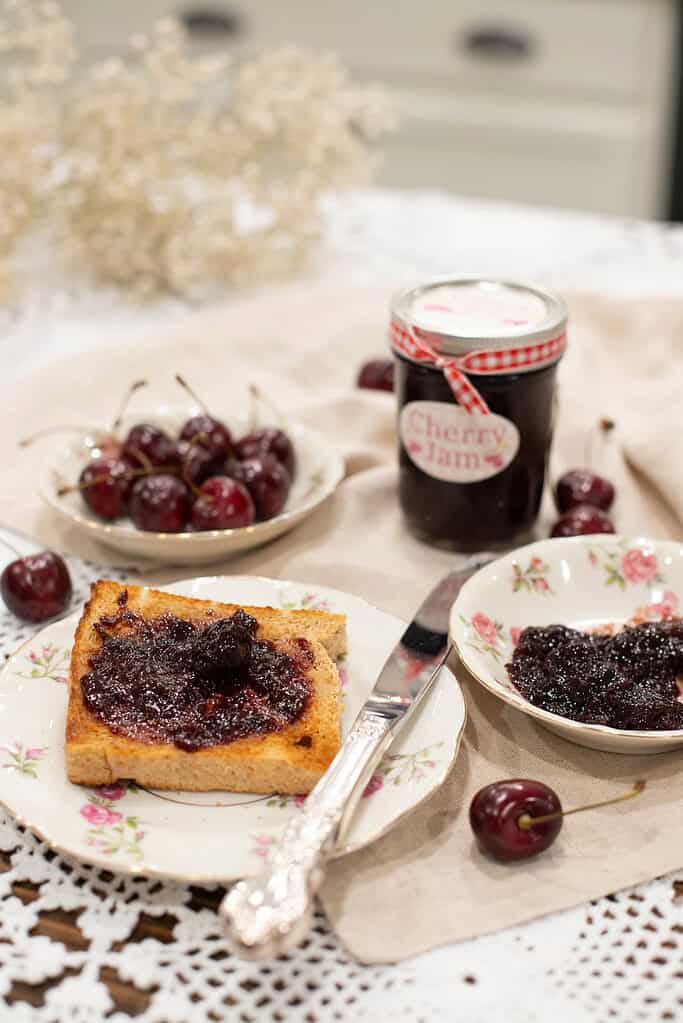 A toasted slice of bread topped with cherry jam, with a whole cherry resting on top, surrounded by vintage dishware and a jar of jam.