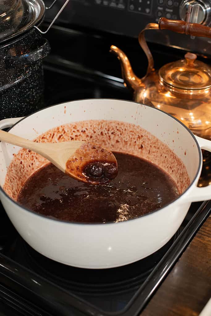 A wooden spoon scooping cooked cherry jam from a white Dutch oven, showing the thick consistency of the homemade preserve.