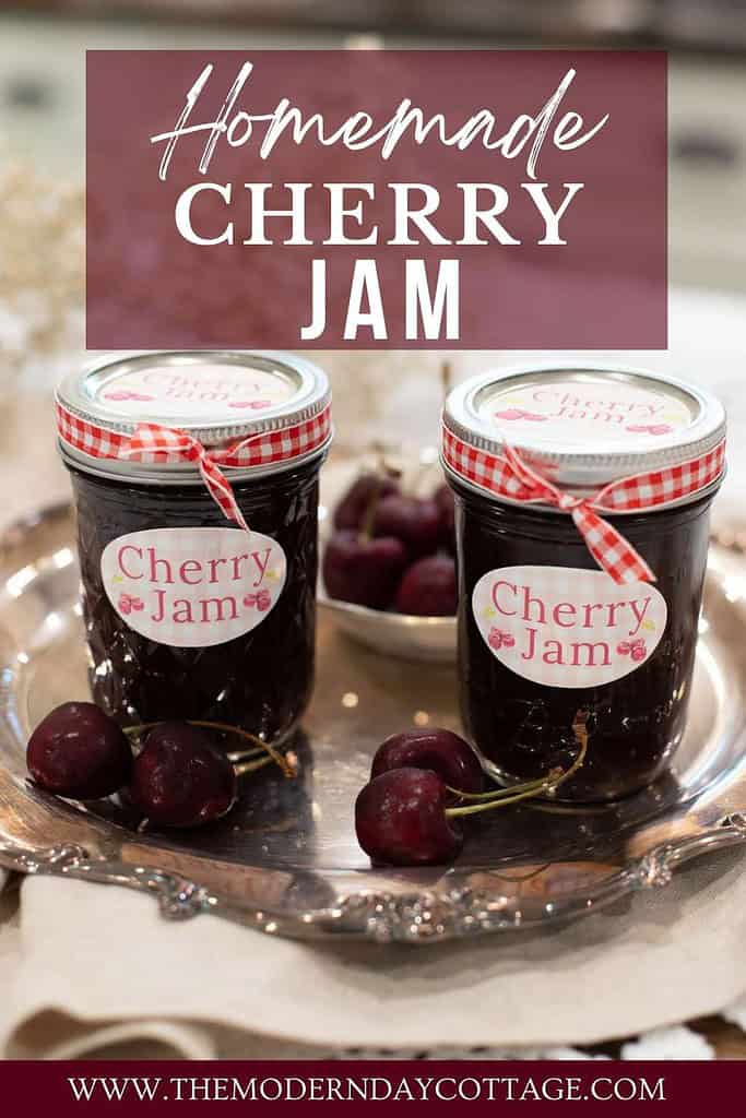 A Pinterest-style graphic featuring two jars of homemade cherry jam tied with red and white gingham ribbon, sitting on a vintage silver tray with fresh cherries. The text overlay reads “Homemade Cherry Jam” in a handwritten and bold serif font, with the website “www.themoderndaycottage.com” displayed at the bottom. The background has a soft, blurred kitchen setting with warm, rustic tones.