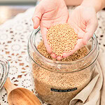 Cupped hands holding hard white wheat berries above a glass jar filled with grain, with a wooden scoop resting on a lace-topped table
